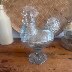 May include: A clear glass rooster-shaped dish with a lid, featuring a textured comb, wattle, and tail feathers. The body of the rooster sits atop a pedestal base with a circular foot, showcasing detailed molded glass patterns throughout the entire piece.