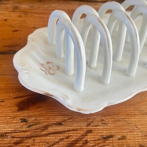 May include: White ceramic toast rack with arched dividers, set on a scalloped base. The rack has a gold trim and a floral design. A charming addition to any breakfast table.