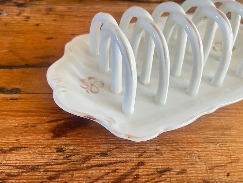 May include: White ceramic toast rack with arched dividers, set on a scalloped base. The rack has a gold trim and a floral design. A charming addition to any breakfast table.