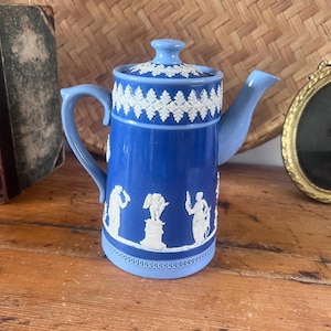 May include: A cylindrical blue teapot featuring white relief figures of classical individuals and a winged cherub on a pedestal. The lid and base are accented with a light blue band and white decorative border patterns, including a spout and handle.
