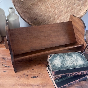 May include: A wooden book stand with a curved top and a shelf, holding two antique books with green covers and black spines. The stand is made of dark wood and sits on a wooden surface. Several antique bottles are in the background.