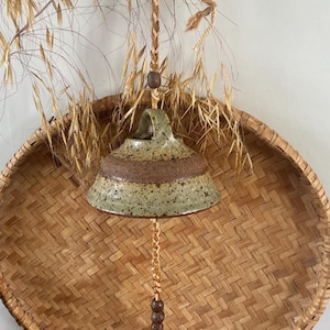 May include: A handcrafted ceramic bell with a brown and green glaze, suspended by a woven rope and wooden beads. The bell hangs in front of a woven basket and dried grasses, creating a rustic, natural aesthetic.