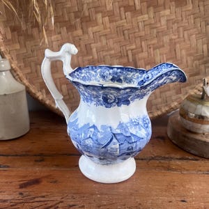 May include: A white and blue ceramic pitcher with a detailed landscape design. The pitcher has a curved spout and a decorative handle. The design features a village scene. The pitcher is on a wooden surface.