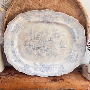 Peut inclure: Un plat ovale en céramique à bord festonné, orné d'un motif de transfert bleu et blanc représentant des bouquets de fleurs et un oiseau. Le fond crème présente de légères taches brunes et le bord affiche un délicat motif en treillis.