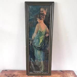 May include: A rectangular framed print featuring a couple in formal attire. A person in a light green beaded dress with a black shawl stands in front of a person in a dark suit. The text "Honeymooning in the Alps" is printed at the bottom.