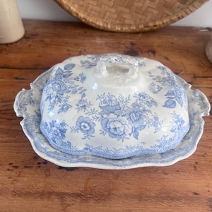May include: A ceramic butter dish with a lid, featuring a cream-colored base and a blue floral pattern. The rectangular dish has scalloped edges and a matching lid with a small handle on top, decorated with intricate blue botanical illustrations.