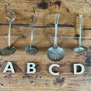 May include: Four antique silver serving spoons with decorative shell and leaf designs are displayed on a wooden surface. Each spoon has a unique handle and bowl shape. Letters A, B, C, and D are placed below each spoon.