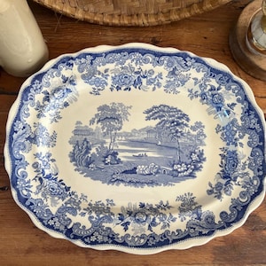 May include: An oval serving platter with a blue and white landscape design. The platter has a scalloped edge and floral border. The central image depicts a scenic view with trees, water, and a bridge.