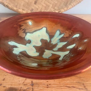 Peut inclure: Un bol rond en céramique émaillée, avec un extérieur brun rougeâtre et un motif intérieur bleu clair et marron. Le bol a une finition brillante et repose sur une surface en bois. Un panier tressé est en arrière-plan.