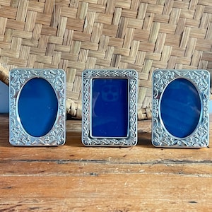 May include: Three silver-colored picture frames with ornate floral designs. Two frames have oval openings, and one has a rectangular opening. Each frame contains a vibrant blue background. The frames are displayed on a wooden surface.