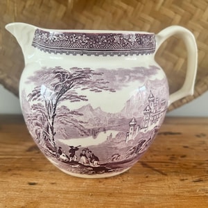 May include: A cream and purple ceramic jug with a landscape design. The jug features a detailed scene of a castle, trees, and figures. The handle and spout are cream, and the jug sits on a wooden surface.