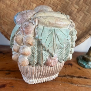 May include: A carved wooden fruit basket sculpture. The sculpture features grapes, leaves, and other fruits in shades of beige, pink, and green. The basket has a textured, ribbed design. The background includes a woven basket and a brass candlestick.
