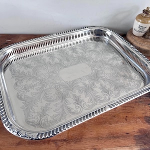 May include: A rectangular silver-toned serving tray with a polished finish, featuring an intricate engraved floral scroll pattern across the centre and a decorative pierced gallery border with a scalloped edge and a smooth central rectangular area.