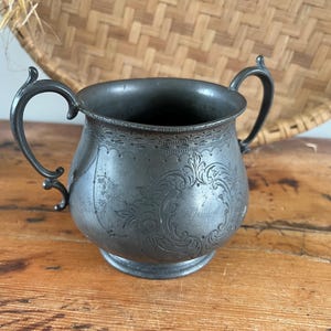 May include: Antique silver-toned sugar bowl with two handles and ornate floral detailing. The vintage bowl sits on a wooden surface, showcasing its aged patina. A decorative piece for serving sugar or other items.