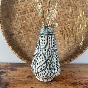 May include: A ceramic vase with a cream base and a blue-grey rim. The vase has a repeating pattern of dark blue lines and dots. Dried flowers are placed inside the vase. The background includes a woven basket and a wooden surface.