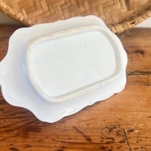 May include: A white ceramic butter dish with a scalloped edge, resting on a matching plate. The dish is rectangular with rounded corners. The set is placed on a wooden surface, with a woven basket in the background.