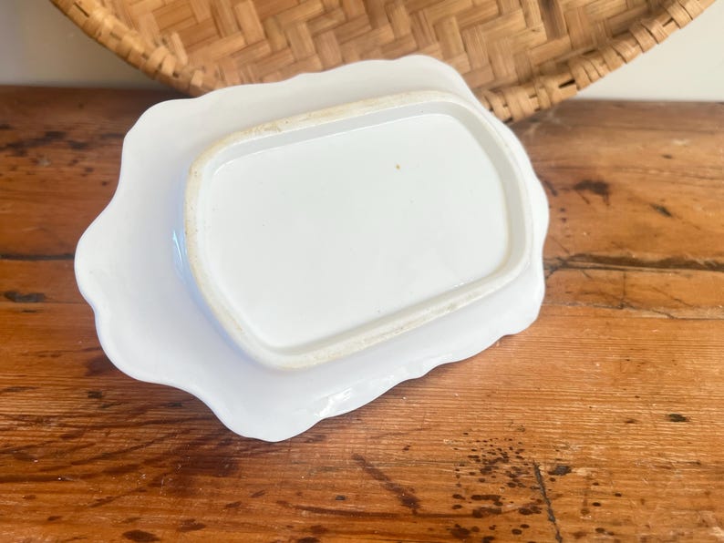 May include: A white ceramic butter dish with a scalloped edge, resting on a matching plate. The dish is rectangular with rounded corners. The set is placed on a wooden surface, with a woven basket in the background.