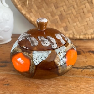 May include: A ceramic lidded jar with a brown glaze, accented with orange and white patterns. The jar has a rounded shape and a small knob on the lid. The design is reminiscent of mid-century modern pottery.