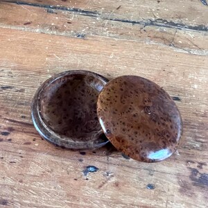May include: A round wooden box with a polished finish and a distinct dark brown burl wood grain pattern. The container consists of a circular base and a separate matching lid, both featuring a smooth, glossy surface with natural wood markings.