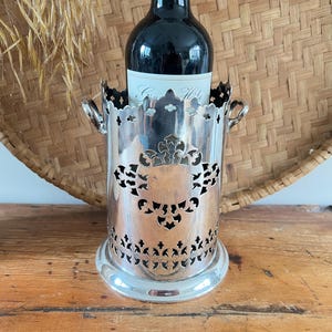 May include: A silver wine bottle holder with a decorative cut-out design. The holder has a round base and two small handles. A bottle of red wine is inside the holder, with a label visible. The background includes a woven basket and a wooden surface.