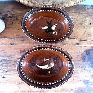 Peut inclure: Deux plats ovales en céramique marron avec des motifs peints à la main. L'un représente un oiseau noir aux ailes blanches, l'autre un poisson blanc. Les deux ont une bordure pointillée noire et blanche.