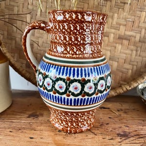 May include: A ceramic jug with a handle, displaying a brown speckled pattern on the top and bottom. The central section has a blue and white floral design with green accents. The jug is on a wooden surface.