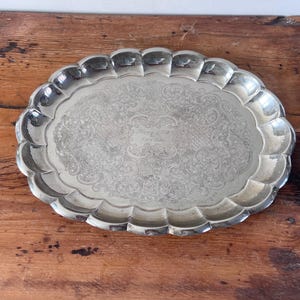 May include: An oval silver-toned metal serving tray featuring a scalloped edge and an intricate engraved floral pattern across the centre surface. The tray has a polished finish and a central rectangular area with faint engraved script text.