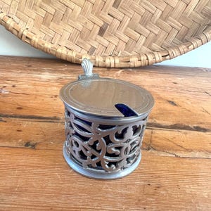 May include: A silver-coloured, ornate container with a lid and a blue interior. The container has a decorative, openwork design around the sides. A small, blue spoon rests inside. The container sits on a wooden surface.
