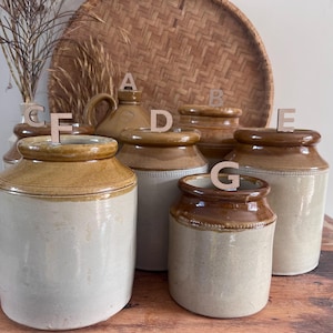 May include: A collection of seven cylindrical stoneware storage jars with two-tone glaze, featuring a light cream-coloured base and a darker brown rim. Each jar is labelled with a small wooden letter tag from A to G for identification purposes.