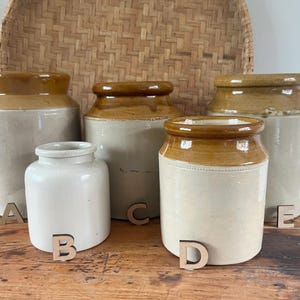 May include: A selection of antique stoneware jars in various sizes and shapes, with tan and cream colour schemes. The jars have a rustic, vintage aesthetic, perfect for kitchen or home decor. The jars are labelled A, B, C, D, and E.