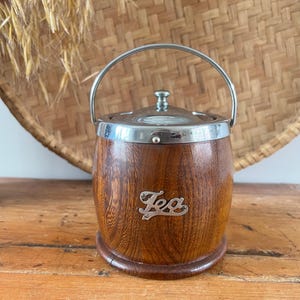 May include: A vintage tea caddy with a wooden barrel-shaped body and a silver-toned lid and handle. The word "Tea" is displayed in a silver script on the front. The caddy sits on a wooden base.
