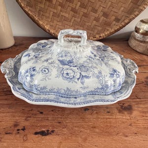 May include: A white ceramic serving dish with a lid, featuring a blue floral pattern. The dish has a scalloped edge and a central handle on the lid. The design includes intricate botanical illustrations of flowers and leaves across the surface.