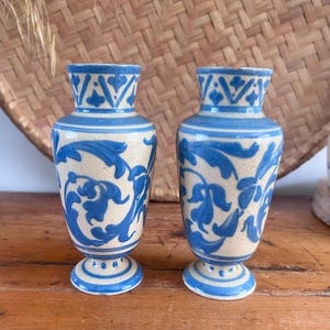 May include: A pair of matching ceramic vases featuring a cream-colored base with hand-painted blue floral scrollwork patterns. Each vase has a flared rim, a slender neck, a bulbous body, and a pedestal base with a small dot design around the edge.