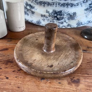 May include: A circular wooden butter press with a central cylindrical handle. The wood features a natural light brown grain with visible signs of wear, including small dark spots and a smooth, rounded edge along the perimeter of the flat base.