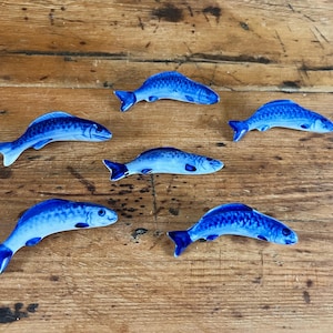 May include: Six blue ceramic fish ornaments with detailed scales and fins are arranged on a wooden surface. The fish vary slightly in size and pose, showcasing a decorative aquatic theme. The ornaments are a vibrant shade of blue.