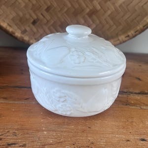 May include: A round white ceramic lidded jar featuring a raised embossed pattern of pears, flowers, and wheat stalks on both the body and the lid. The lid includes a small, smooth, rounded knob handle at the center for easy removal and placement.