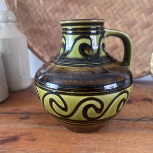 May include: A ceramic pitcher with a rounded body and a single handle, featuring a glossy glaze in shades of olive green and dark brown. The surface is decorated with a repeating dark brown wave pattern against a light olive green background.