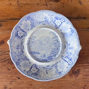 May include: A round ceramic plate featuring a blue and white transferware pattern. The centre depicts a landscape with figures and buildings, while the scalloped rim is decorated with detailed floral motifs and intricate scrollwork.