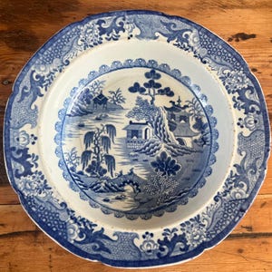 May include: A round, antique blue and white ceramic plate with a scalloped edge. The plate features a detailed landscape scene with buildings, trees, and a bridge. The border has floral and scrollwork designs.