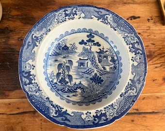 May include: A round, antique blue and white ceramic plate with a scalloped edge. The plate features a detailed landscape scene with buildings, trees, and a bridge. The border has floral and scrollwork designs.