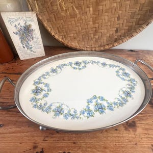 May include: Oval serving tray with a white centre and silver-coloured rim and handles. The tray features a floral design in shades of blue. The tray is on a wooden surface.