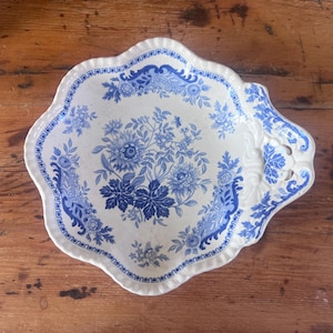 May include: A white and blue ceramic serving platter with a floral design. The platter has a scalloped edge and handles. The floral pattern features blue flowers and leaves on a white background. The platter is likely vintage or antique.
