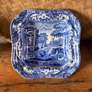 May include: A square ceramic dish with rounded corners featuring a blue and white transferware pattern. The center depicts a landscape with ruins, trees, and figures, while the wide border displays a detailed floral and scrollwork design.
