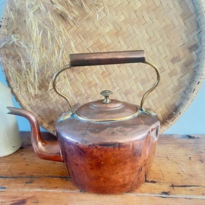 May include: A copper kettle with brass detailing and a wooden handle. The kettle has a rounded body, a curved spout, and a lid with a small knob. It is displayed on a wooden surface, with a woven basket and a white vase in the background.