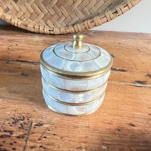 May include: A small, round decorative box with a lid. The box is covered in iridescent, mother-of-pearl tiles and trimmed with gold-colored metal. The lid has a small, round gold-colored knob. The box sits on a wooden surface.