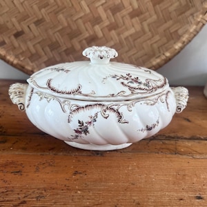 May include: A white ceramic tureen with a swirling ribbed texture and a matching lid. The piece features brown decorative scrollwork and floral patterns along the rim and body, complemented by ornate handles and a central knob on the lid.