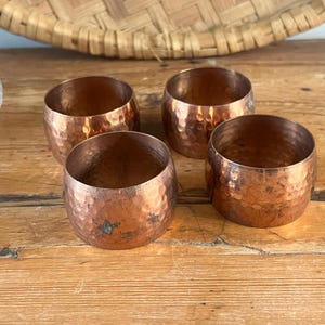 May include: Four copper-coloured metal napkin rings with a hammered texture. The rings are cylindrical and sit on a wooden surface. A woven basket and a glass dish are in the background.
