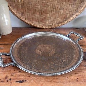 May include: A round silver-toned serving tray featuring an intricate embossed floral pattern across its surface. The tray includes two ornate, textured handles on opposite sides and a decorative rope-style border along the outer circular rim.
