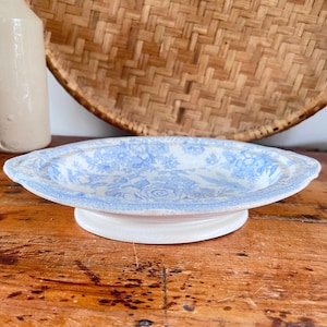 May include: An oval ceramic dish featuring a light blue floral pattern on a white background. The dish has a raised pedestal base and small handles on each side, with a detailed transferware design covering the interior surface.
