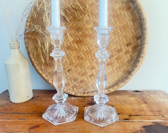 May include: Two clear glass candle holders with white candles. The candle holders have a faceted design and sit on a wooden surface. A woven basket and a ceramic jug are in the background.